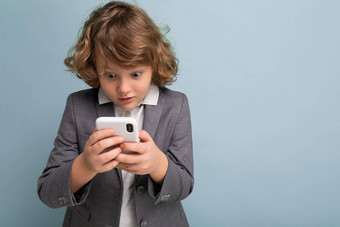 照片英俊的震惊了男孩卷曲的头发穿灰色西装持有电话孤立的蓝色的背景智能手机沟通生活短信