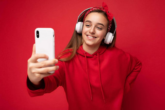 特写镜头拍摄微笑漂亮的浅黑肤色的女人女孩穿红色的连帽衫孤立的红色的背景持有智能手机采取自拍照片穿白色无线耳机听很酷的音乐电话显示