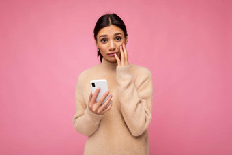 有吸引力的可爱的年轻的心烦意乱伤心褐色女人穿米色温暖的毛衣站孤立的粉红色的背景冲浪互联网电话相机坏情绪