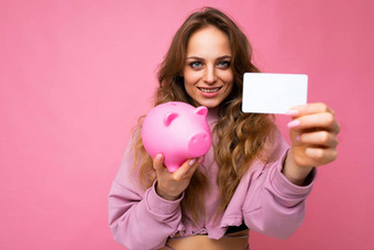 快乐积极的微笑年轻的美丽的迷人的女人波浪长光头发真诚的情绪穿时尚的粉红色的连帽衫孤立的粉红色的背景复制空间持有粉红色的小猪盒子信贷卡银行概念