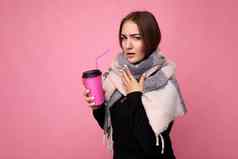 照片拍摄美丽的年轻的浅黑肤色的女人女人穿黑色的毛衣温暖的围巾孤立的粉红色的背景持有纸咖啡杯模型喝热饮料冷