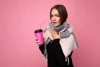 照片拍摄美丽的年轻的浅黑肤色的女人女人穿黑色的毛衣温暖的围巾孤立的粉红色的背景持有纸咖啡杯模型喝<strong>热饮料</strong>冷