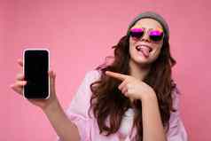 积极的有吸引力的年轻的浅黑肤色的女人女人穿时尚的粉红色的衬衫灰色他色彩鲜艳的太阳镜孤立的粉红色的背景持有手显示移动电话空显示模型相机指出手指gadjet显示舌头
