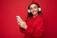 特写镜头肖像照片美丽的积极的微笑褐色女少年穿红色的连帽衫孤立的红色的背景持有智能手机沟通在线穿白色无线耳机听很酷的音乐相机