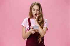 女孩发现令人震惊的真理阅读消息智能手机紧迫的手臂脸颊惊喜盯着有关说不出话来手机屏幕反应意想不到的新闻粉红色的墙