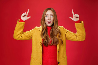 惊讶红色头发的人女孩黄色的外套开放口深刻的印象激动提高手指出告诉很酷的促销活动兴奋表达式红色的背景