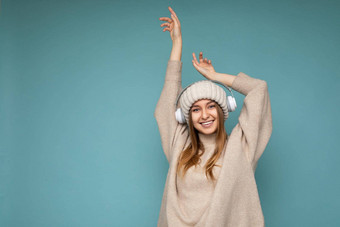 有吸引力的积极的微笑年轻的金发女郎女人穿米色冬天毛衣他孤立的蓝色的背景墙穿白色无线蓝牙耳机听音乐有趣的相机