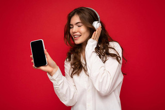 有吸引力的积极的年轻的浅黑肤色的女人卷曲的女人穿白色衬衫孤立的红色的背景墙持有显示移动电话空显示穿白色蓝牙耳机听音乐gadjet