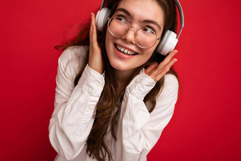 特写镜头照片有吸引力的积极的微笑年轻的褐色女人穿白色衬衫光学眼镜孤立的红色的背景穿白色无线蓝牙耳机听音乐有趣的一边
