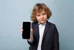 特写镜头肖像英俊的积极的男孩卷曲的头发穿西装持有电话孤立的蓝色的背景相机显示智能手机空显示屏幕