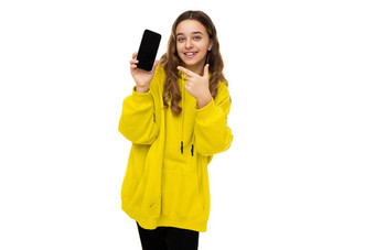 照片美丽的可爱的微笑积极的有趣的浅黑肤色的女人十几岁的女孩时尚的黄色的体育连帽衫持有移动电话黑色的空白屏幕孤立的白色背景免费的空间文本