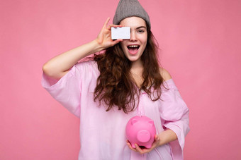 迷人的快乐快乐的年轻的浅黑肤色的女人女人穿衬衫孤立的粉红色的背景<strong>免费</strong>的空间持有粉红色的猪钱盒子信贷卡<strong>模型</strong>