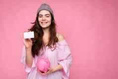肖像美丽的积极的快乐的可爱的微笑年轻的浅黑肤色的女人女人时尚的衬衫孤立的粉红色的背景复制空间持有粉红色的猪Moneybox信贷卡模型