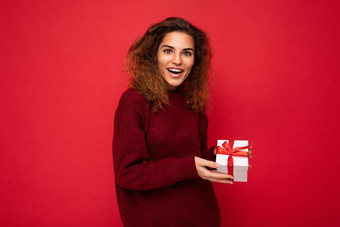 有吸引力的积极的惊讶年轻的浅黑肤色的女人卷曲的女人孤立的红色的背景墙穿红色的毛衣持有礼物盒子相机