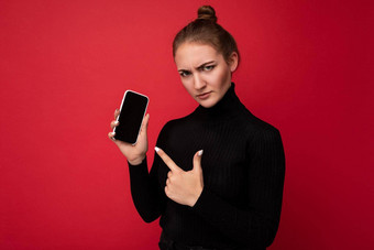 愤怒的伤心有吸引力的积极的年轻的浅黑<strong>肤色</strong>的女人女人穿黑色的毛衣站孤立的红色的背景显示移动电话空屏幕模型相机指出<strong>手指</strong>gadjet