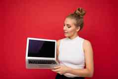 照片美丽的金发碧眼的年轻的女人聚集头发上网本键盘持有电脑移动PC空监控屏幕模拟复制空间穿白色t恤孤立的红色的墙背景