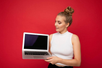 <strong>照片</strong>美丽的金发碧眼的年轻的女人聚集头发上网本键盘持有电脑移动PC空监控屏幕模拟复制空间穿白色t恤孤立的红色的<strong>墙背景</strong>