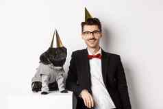 图像英俊的年轻的男人。庆祝生日可爱的黑色的哈巴狗聚会，派对服装锥头站白色背景