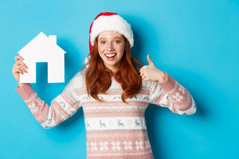 假期广告片真正的房地产概念满意女模型红色的波浪头发穿圣诞老人他毛衣显示纸房子模型竖起大拇指蓝色的背景