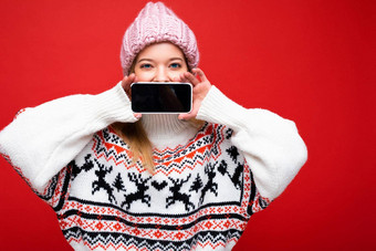 照片有吸引力的快乐年轻的金发女郎女人穿<strong>温暖</strong>的针织他冬天<strong>温暖</strong>的毛衣站孤立的红色的背景显示智能手机空屏幕模拟设备显示