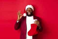 惊讶黑色的男人。持有假期礼物内部圣诞节袜子显示标志批准站红色的背景