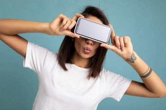 照片漂亮的积极的年轻的浅黑肤色的女人女人好穿白色t恤站孤立的蓝色的背景复制空间持有智能手机显示电话手空屏幕屏幕模型给吻