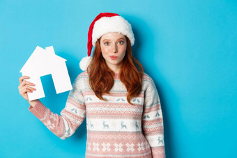 假期广告片真正的房地产概念可爱的红色头发的人女人圣诞老人他毛衣显示纸房子模型公寓提供站蓝色的背景