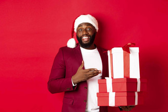 圣诞节一年购物概念快乐的黑色的男人。秘密圣诞老人持有圣诞节礼物微笑兴奋带礼物站红色的背景