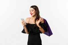 美购物概念华丽的女人优雅的黑色的衣服化妆订单在线智能手机持有袋微笑站白色背景