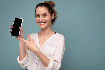 美丽的积极的年轻的金发女郎卷曲的女人穿休闲白色衬衫孤立的蓝色的背景墙持有电话显示智能手机空dcreen模型相机指出手指设备