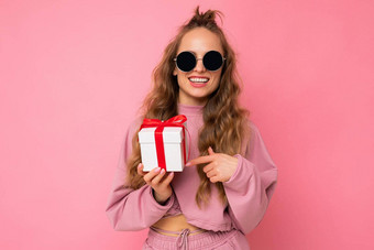 有吸引力的快乐微笑年轻的金发女郎卷曲的女人孤立的粉红色的背景墙穿粉红色的体育运动衣服太阳镜持有礼物盒子相机