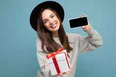 拍摄漂亮的微笑积极的年轻的褐色女人孤立的蓝色的背景墙穿时尚的黑色的他灰色毛衣持有礼物盒子显示智能手机屏幕显示模型相机