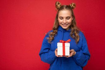 拍摄有吸引力的快乐微笑年轻的金发女郎女人孤立的红色的背景墙穿蓝色的时尚的连帽衫持有礼物盒子白色盒子红色的丝带