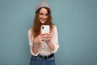 照片有吸引力的积极的年轻的金发女郎卷曲的女人穿休闲白色衬衫灰色他孤立的蓝色的背景墙持有电话沟通在线相机