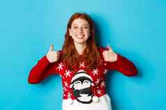 冬天假期圣诞节夏娃概念快乐微笑女孩红色的头发显示拇指批准赞美好产品推荐站圣诞节毛衣