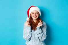 冬天假期圣诞节夏娃概念充满希望的红色头发的人女孩圣诞老人他使圣诞节手指交叉穿圣诞老人他站蓝色的背景