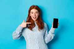 深刻的印象红色头发的人女孩指出电话屏幕显示智能手机应用程序在线提供微笑兴奋站毛衣蓝色的背景
