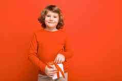 积极的微笑金发女郎卷曲的男孩孤立的红色的背景墙穿红色的毛衣持有礼物盒子相机