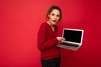 特写镜头肖像美丽的惊讶惊讶惊讶黑暗金发碧眼的女人持有移动PC电脑相机显示上网本键盘穿红色的毛衣孤立的红色的墙背景