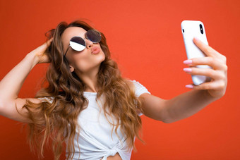 令人惊异的美丽的年轻的<strong>女人</strong>持有移动电话采取自<strong>拍照片</strong>智能手机相机穿太阳镜日常时尚的装孤立的色彩斑斓的墙背景设备屏幕