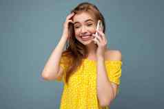 有吸引力的积极的微笑年轻的金发女郎女人穿时尚的黄色的夏天衣服站孤立的蓝色的背景持有会说话的移动电话一边