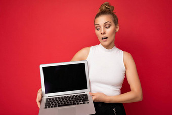 美丽的金发碧眼的年轻的女人聚集头发上网本键盘持有电脑移动PC空监控屏幕模拟复制空间穿白色t恤孤立的红色的墙背景