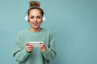 特写镜头照片美丽的积极的快乐年轻的女人穿蓝色的毛衣孤立的蓝色的背景穿白色蓝牙无线耳机听音乐移动电话沟通在线互联网相机