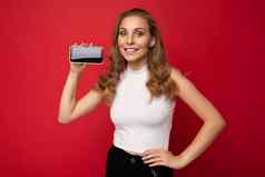 微笑美丽的年轻的金发女郎女人穿白色t恤孤立的红色的背景复制空间持有智能手机显示电话手空屏幕模型相机