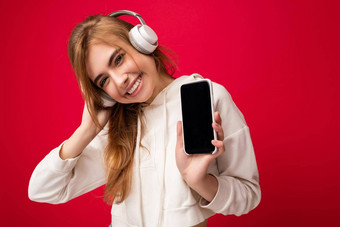 美丽的快乐微笑年轻的女人穿时尚的休闲装孤立的背景墙持有显示移动电话空显示模型穿白色<strong>蓝牙耳机</strong>听音乐有趣的相机