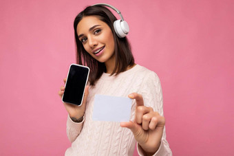 有吸引力的积极的微笑年轻的女人穿时尚的休闲装孤立的色彩鲜艳的背景墙持有信贷卡显示移动电话空屏幕断路穿白色蓝牙耳机有趣的相机