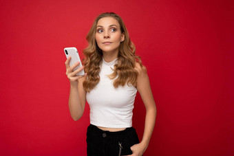 快乐漂亮的年轻的金发女郎女人穿白色t恤孤立的红色的背景智能手机聊天移动电话一边