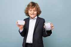 英俊的积极的快乐男孩卷曲的头发穿西装持有电话信贷卡显示移动屏幕相机孤立的蓝色的背景塑料卡