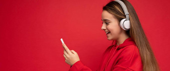 全景侧面轮廓拍摄积极的微笑漂亮的浅黑肤色的女人女孩穿红色的连帽衫孤立的红色的背景持有智能手机写作短信消息穿白色无线耳机听很酷的音乐电话显示