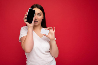有吸引力的快乐年轻的浅黑肤色的女人女人穿休闲白色t恤孤立的红色的背景空空间持有手移动电话显示智能手机空屏幕模型信贷卡相机